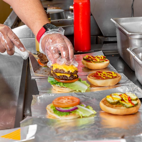 Gloved hands assemble Home Run Burger's double cheeseburgers with fresh toppings on a foil-lined counter.