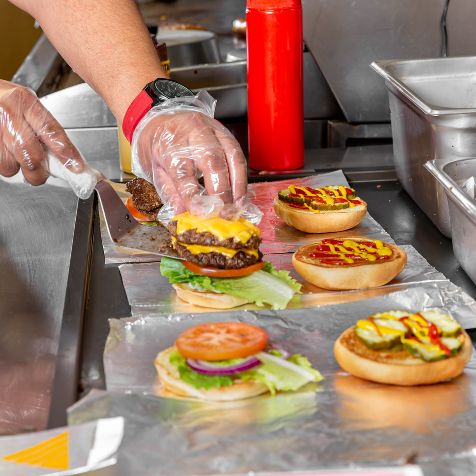 Gloved hands assemble Home Run Burger's double cheeseburgers with fresh toppings on a foil-lined counter.