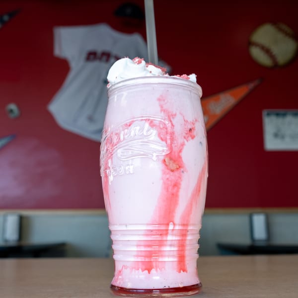 A strawberry milkshake with whipped cream and sprinkles in a mason jar, against a red wall.