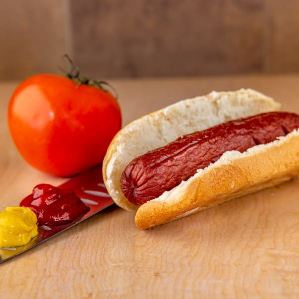 A hot dog in a bun with a tomato, ketchup, and mustard on a wooden surface.