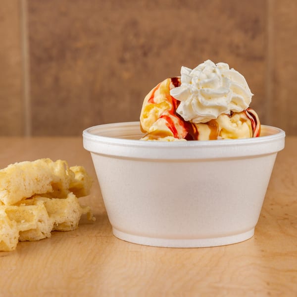 Vanilla ice cream with strawberry and caramel drizzle, topped with whipped cream, next to waffle pieces.