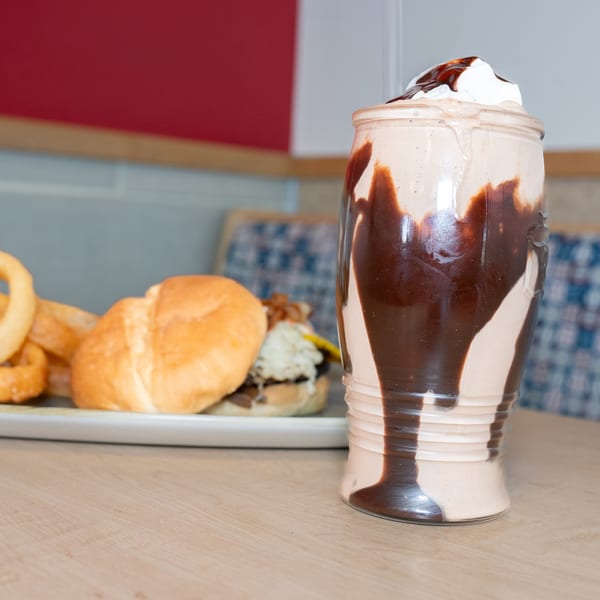 Chocolate milkshake with whipped cream and chocolate drizzle, with a burger and onion rings in the background.