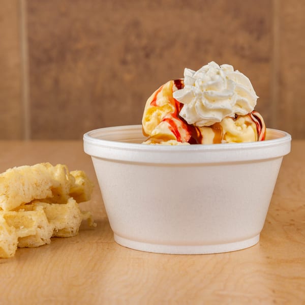 Vanilla ice cream with strawberry and caramel syrup, topped with whipped cream, next to waffle pieces.