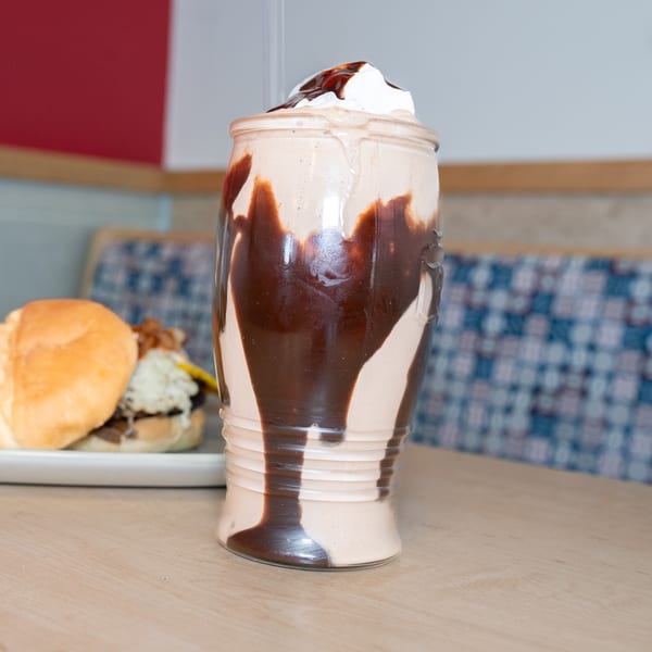A chocolate milkshake with whipped cream and chocolate drizzle, with a burger in the background.