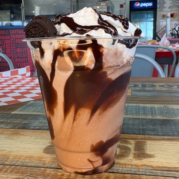 A chocolate milkshake with whipped cream, chocolate drizzle, and an Oreo cookie on a wooden table.